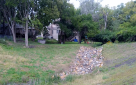 Photo of rip rap along new drainage ditch surrounded by green grass, trees and behind homes