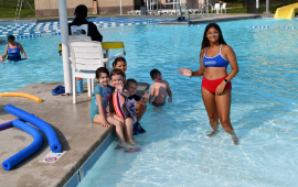 Woman in a bathing suit smiling and waving in a pool with a group of children