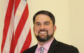 Image of man standing next to an American flag wearing a long necktie and black blazer