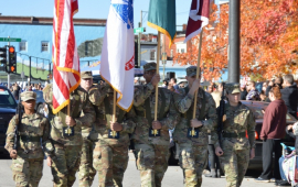Honor Guard dressed in Army uniforms carry flags on a sunny street with orange trees turned for fall.