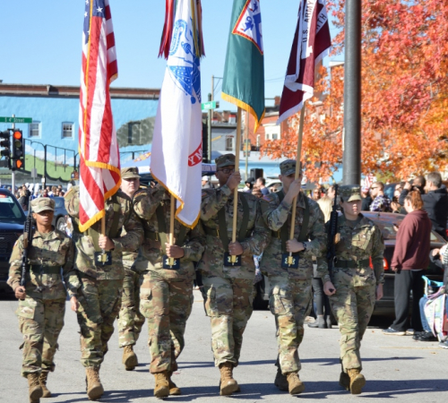 Honor Guard dressed in Army uniforms carry flags on a sunny street with orange trees turned for fall.