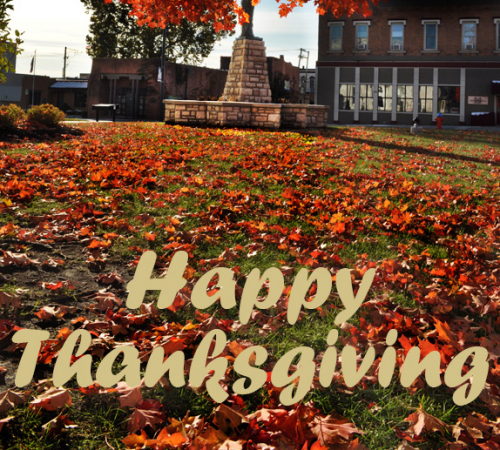 Tree with orange leaves, some on the ground, around mini Statue of Liberty, Leavenworth city logo text reads Happy Thanksgiving