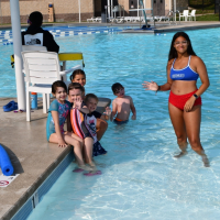 Woman in a bathing suit smiling and waving in a pool with a group of children