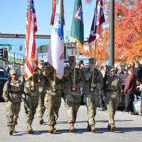 Honor Guard dressed in Army uniforms carry flags on a sunny street with orange trees turned for fall.
